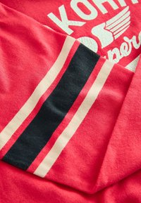 Rød T-shirt med hvidt, revnet tekst og et vinge-motiv. Tykkere marineblå og elfenbensstriber fremhæver ærmerne, og stoffet er blødt bomuld.