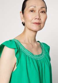 Blusa verde com um design evasé, apresentando um decote em forma de onda adornado com bordados brancos. Tecido suave e leve com uma textura lisa.