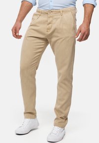 Des chinos beiges en coton présentent une coupe ajustée, des plis à l'avant et une fermeture à bouton. Portés avec des baskets blanches.