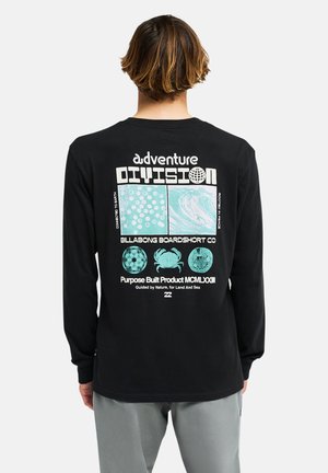 Persona che indossa una maglietta nera a maniche lunghe con grafiche turchesi a tema natura e testo "adventure division" sulla schiena su sfondo bianco.