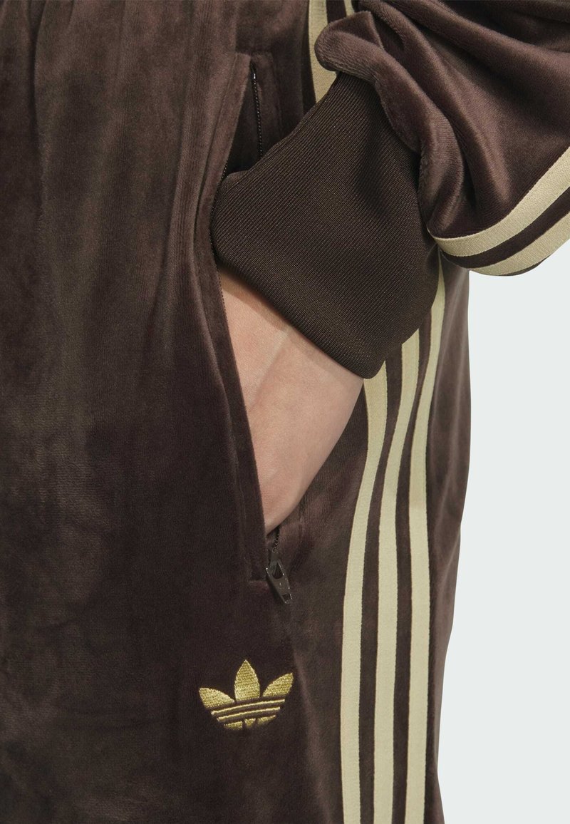 Hand in de zak van een bruine velours jas met beige strepen en een gouden Adidas-logo aan de onderkant.