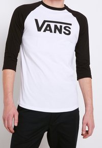 Schwarzes und weißes Vans T-Shirt mit schwarzem Körper und Ärmeln, versehen mit einem auffälligen schwarzen Logo auf der Brust. Aus Baumwollstoff.