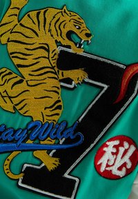 Un tessuto verde presenta un tigre ricamato in giallo e nero, accanto a un "7" nero e a una scritta blu "Stay Wild", con un emblema circolare rosso.