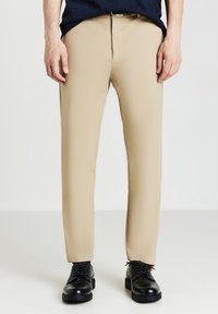 Beige Maßhose mit glatter Textur, die einen geraden Schnitt und einen flachen Bund aufweist. Kombiniert mit schwarzen Schnürschuhen.