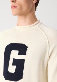 Cream-colored knit sweater featuring a large navy blue "G" logo, ribbed crew neckline, and textured raglan sleeves.