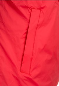 Red waterproof jacket material with a smooth texture, featuring a hidden side pocket sewn into the midsection. Seam details visible.