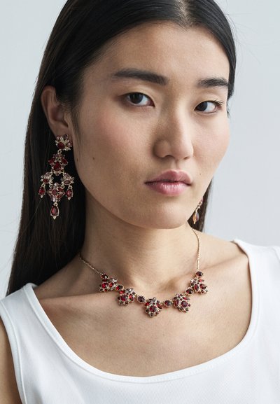 Náhrdelník s červeným drahokamem a odpovídajícími křišťálovými náušnicemi se zlatými akcenty, které mají složité květinové vzory a facets. 