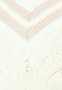 Neulottu viltti, jossa on chevron-kuvio, vaihtuvilla raitoja vaaleassa ja vaaleanbeigeissä väreissä, pehmeällä ja pörröisellä pinnalla.