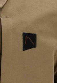 Veste beige en tissu texturé ; dotée d'un patch noir avec un motif brodé orange sur la poitrine. Lignes épurées et détails minimalistes.