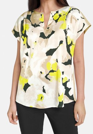 Femme portant un chemisier ample à motifs floraux beiges, jaunes, blancs et noirs abstraits et un pantalon noir, bras détendus le long du corps.