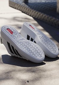 Baskets compensées blanches avec des rayures noires et des semelles à motifs, posées semelle vers le haut sur une surface en béton ensoleillée près d'une corde.
