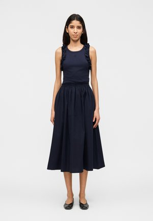 Naine seisab näoga otse, kandes varrukateta tumelõhnavat topi volangidega ja vastavat tumelõhnavat midi-seelikut mustade tasaste jalanõudega.