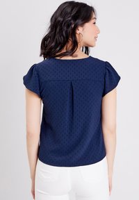 Haut bleu marine avec des manches courtes bouffantes, tissu texturé présentant un motif subtil et un détail de pli au dos ; porté avec un pantalon blanc.