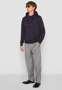 Sweat à capuche bleu marine avec cordon de serrage, poche avant et logo ; associé à un pantalon plissé gris clair et des chaussures noires. Le mannequin porte des lunettes de soleil.