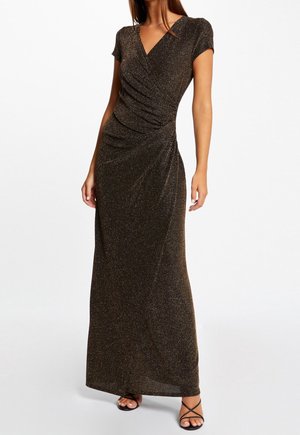 Femme portant une robe portefeuille longue jusqu'au sol, sombre et scintillante, avec des manches courtes et des talons ouverts noirs à brides, debout sur un fond blanc.