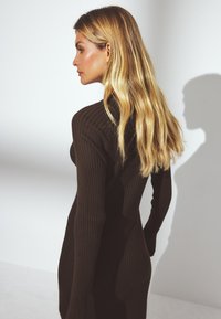 Pikad lokkis juustega blond naine, kes kannab sobivat, pikkade varrukatega, soonikutest tumeda pruuni kleiti, seistes vasakule poole valge tausta ees.