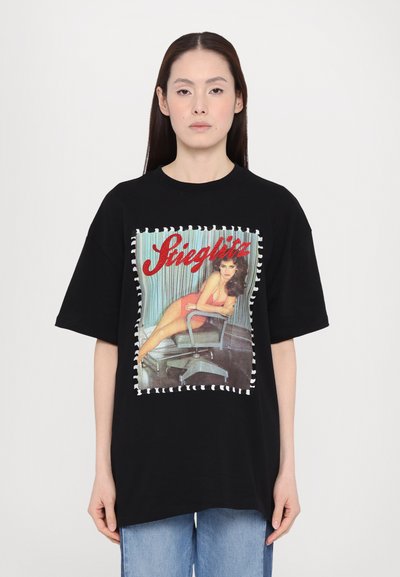 Tricou negru oversized cu un imprimeu grafic vintage, marginit de cusături albe. Designul include o figură așezată și text stilizat.