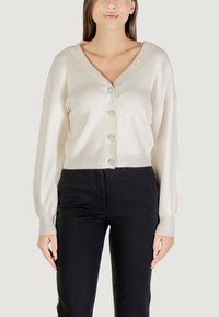Cárdigan de canalé crema con un profundo escote en V, terminado con botones perlados y mangas abullonadas, combinado con pantalones negros. Textura suave.