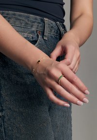 Złota bransoletka z zielonym kamieniem szlachetnym i delikatnym łańcuchem, w komplecie ze złotym pierścionkiem z zielonym kamieniem i przezroczystymi akcentami na dłoni.