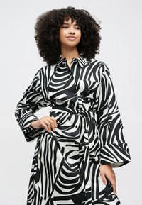 Moteris su banguotais plaukais, dėvinti juoda ir balta dryžuotą zebros rašto su diržu marškinių suknelę, poza viena ranka ant klubo prie paprasto fono.