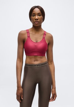 Atletische vrouw staand, gekleed in een rode sportbeha en bruine Under Armour leggings, tegen een effen lichte achtergrond.
