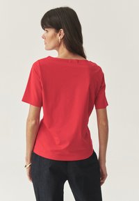 Top scurt roșu cu mâneci scurte, cu decolteu larg și textură moale, asortat cu pantaloni din denim închis. Design simplu, fără modele vizibile sau embellishments.