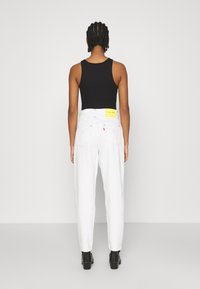 Bílé vysoké džíny s kapsami na zadní straně a žlutou cedulkou Levi's Red. Modelka má na sobě černý top na ramínka a černé boty na podpatku.