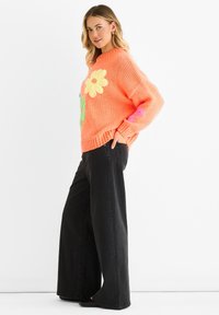 Pomarańczowy sweter dziany z dużymi aplikacjami kwiatowymi w kolorze żółtym, różowym i zielonym; zestawiony z czarnymi spodniami o szerokich nogawkach.