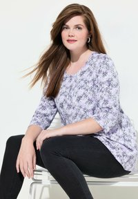 Lys lilla top med et blomsterprint i sort og hvid, 3/4 ærmer, rund halsudskæring, parret med sorte bukser, siddende på en hvid stol.