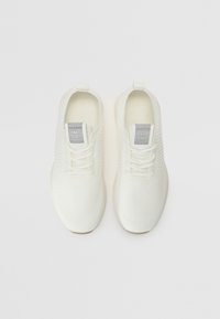 White lace-up sneakers with a textured knit upper, rounded toe, and a grey logo patch on the tongue. Soft, flexible sole.