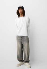 Oversized white long-sleeve shirt, light wash denim jeans with faded texture, paired with cream sneakers and a dark baseball cap.