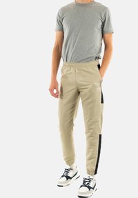 Pantalones deportivos beige con paneles laterales negros, cinturilla elástica y tobillos ajustados. Combinados con una camiseta gris y zapatillas deportivas.