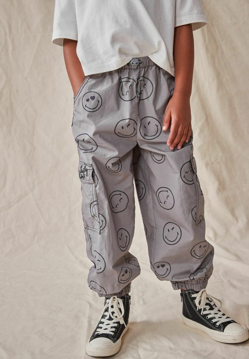 Kind in grauen Hosen mit schwarzen Smileys, weißem T-Shirt und schwarzen High-Top-Sneakers mit weißen Schnürsenkeln vor einem neutralen Hintergrund.