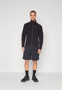 Νεαρός άνδρας στέκεται σε εσωτερικό χώρο, φορώντας μαύρο μπουφάν με φερμουάρ, σκούρο cargo σορτς, μαύρες κάλτσες και μαύρα αθλητικά παπούτσια.