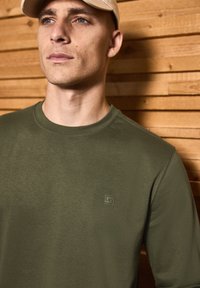 Olivově zelený svetr s kulatým výstřihem vyrobený z měkké tkaniny, s jemným logem na hrudi, na pozadí dřevěného povrchu.