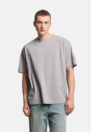 Młody mężczyzna z krótkimi włosami, ubrany w luźny jasno szary t-shirt z krótkim rękawem i jasnoniebieskie dżinsy, stoi na jednolitym tle.