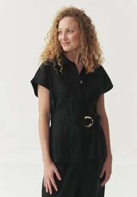 Zwarte shirt met korte mouwen, knoopsluiting aan de voorkant en een brede riem met een ronde gesp. Glad materiaal met een relaxte pasvorm.