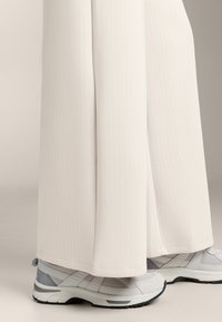 Bijele hlače od rebrastog materijala s širokim nogavicama, uparene sa svijetlosivim i bijelim sportskim cipelama s teksturiranim dizajnom i potpornih đonom.