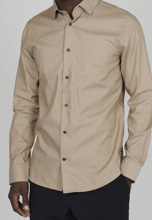 Beige Langarmhemd mit Knopfleiste und strukturiertem Finish, ausgestattet mit einem spitzen Kragen und dunklen Braunen Knöpfen, kombiniert mit dunklen Hosen.