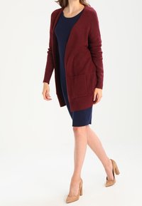 Cardigan bordeaux lavorato a maglia con maniche lunghe e tasca frontale, indossato sopra un vestito blu navy, abbinato a scarpe con tacco nude.