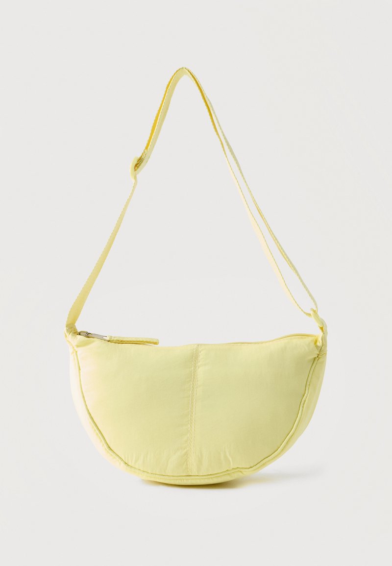 Zachte gele schoudertas in vorm van een halve maan met verstelbare band en ritssluiting bovenaan tegen een effen lichte achtergrond.