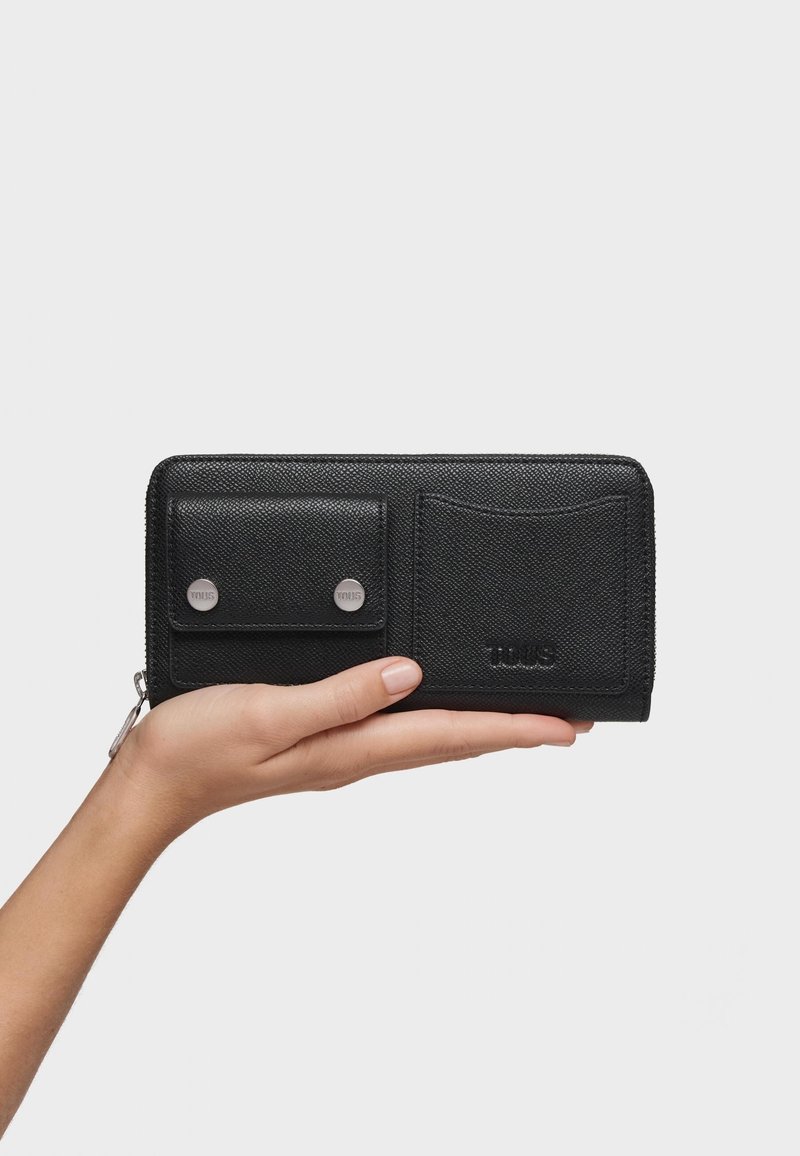 Main tenant un portefeuille en cuir noir avec une poche à monnaie boutonnée et un nom de marque embossé à l'avant sur un fond blanc.