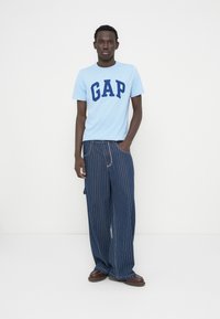 T-shirt bleu clair avec l'inscription « GAP » en lettres bleu marine, associé à un pantalon large à fines rayures bleu marine et des chaussures marron. Tissu en coton lisse.