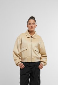 Béžová umelá semišová bomber bunda so zipsom vpredu, veľkými vreckami na klopu, rebrovanými manžetami a lemom. Nosená s čiernymi cargo nohavicami.
