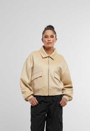 Beige Kunstleder-Bomberjacke mit einem Reißverschluss vorne, großen Klappentaschen, gerippten Bündchen und Saum. Kombiniert mit schwarzen Cargo-Hosen.