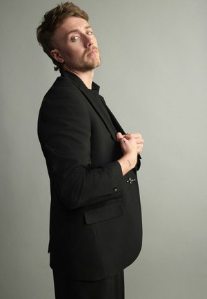 Man with light brown hair wearing black suit jacket, clasping hands near chest, looking slightly upward against plain gray background.