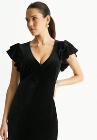 Black velvet dress with a V-neck and flared ruffle sleeves. Smooth texture, form-fitting silhouette, and elegant drape. Gold earrings as an accent.