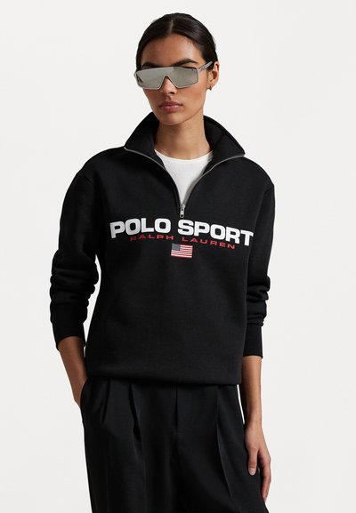 Juodas užtrauktuku užsegamas džemperis su "POLO SPORT" ir vėliavos grafika, pagamintas iš minkštos medžiagos. Dėvimas su veidrodiniais akiniais nuo saulės ir juodomis kelnėmis.