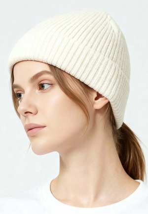 Jeune femme à la peau claire et aux cheveux bruns attachés en queue de cheval basse, portant un bonnet en tricot côtelé blanc et une chemise blanche, regardant vers la gauche.