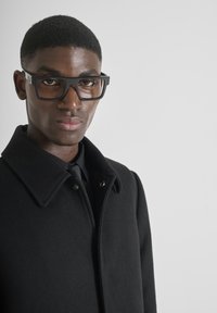 Cappotto di lana nera con colletto classico, dotato di due bottoni a pressione. Grandi occhiali neri rettangolari su sfondo neutro.